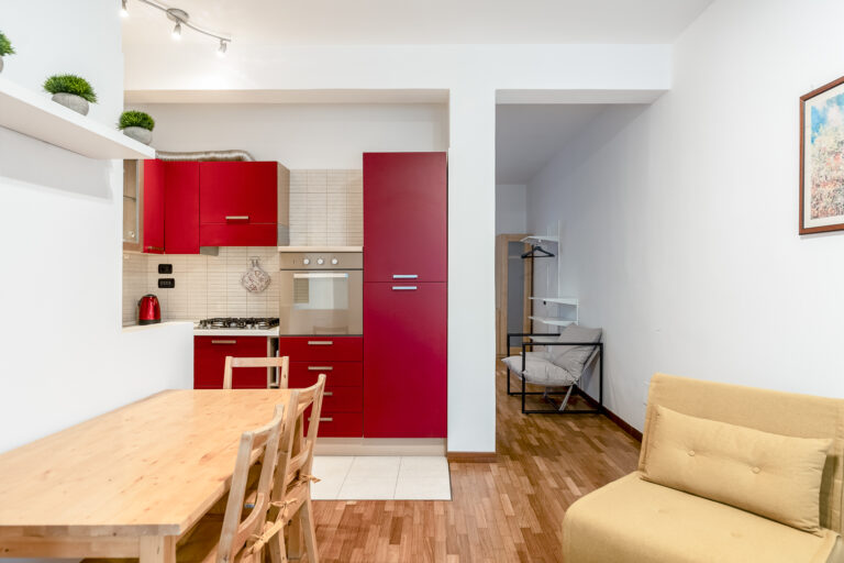 Cocina con gabinetes rojos, mesa de madera, sillas, sofá amarillo y estante con plantas. Área iluminada y moderna.