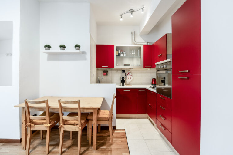 Cocina moderna con muebles rojos, mesa de madera con sillas, horno empotrado, y pequeños maceteros en una repisa.