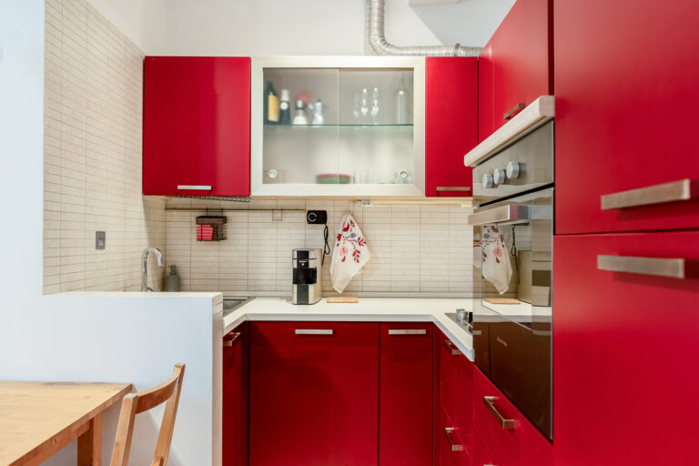 Cocina moderna con muebles rojos, estufa, fregadero de acero, mesa de madera y azulejos blancos. Accesorios y utensilios a la vista.