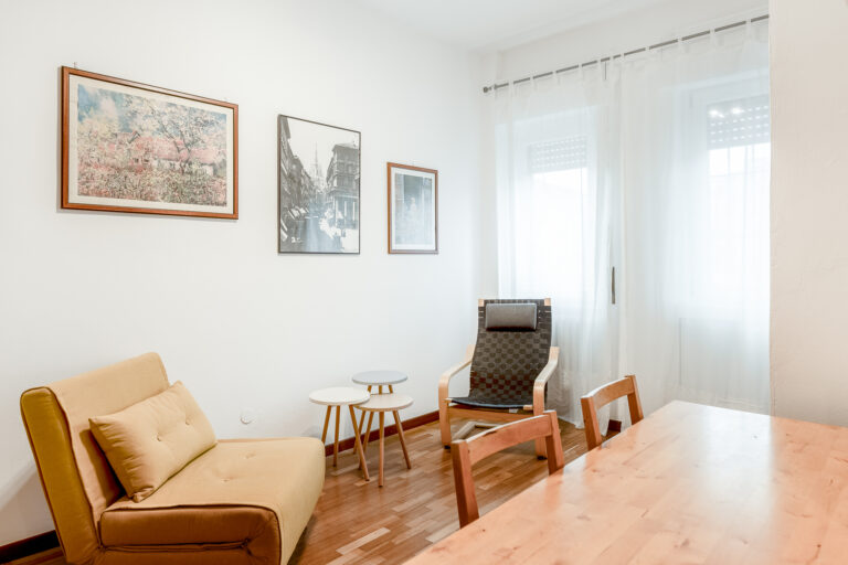 Sala moderna con sillón beige, silla de diseño, mesas auxiliares blancas y cuadros en la pared, iluminada por una ventana con cortinas blancas.