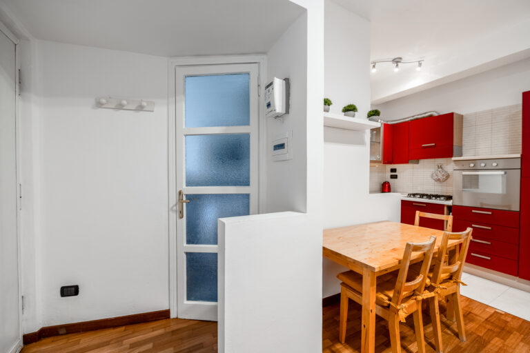 Cocina acogedora, paredes blancas, puerta de cristal opaco, muebles rojos, mesa de madera clara con dos sillas.