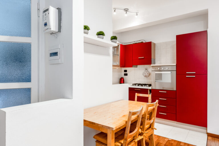 Cocina moderna con gabinetes rojos, mesa de madera clara y sillas. Encimera de gas, horno empotrado y decoración minimalista.