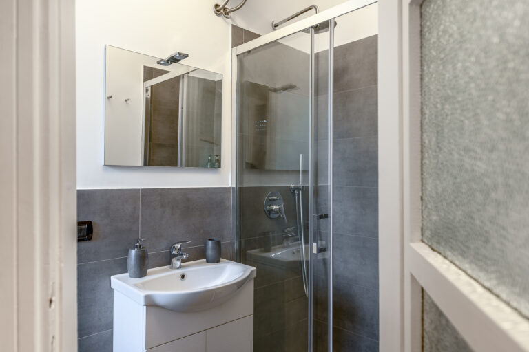 Baño moderno con ducha de vidrio, lavabo blanco, espejo rectangular y paredes grises. Accesorios de baño en gris.
