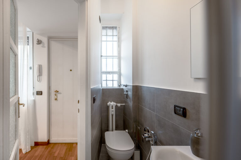 Entrada de un apartamento, puerta blanca, suelo de madera, cuarto de baño pequeño con inodoro, lavabo y azulejos grises.