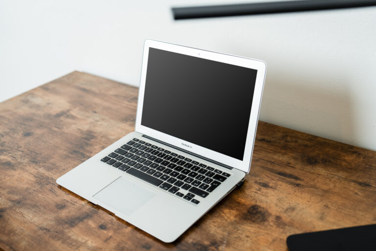 Laptop MacBook Air con pantalla negra sobre mesa de madera desgastada. Fondo de pared blanca y objeto negro al borde.