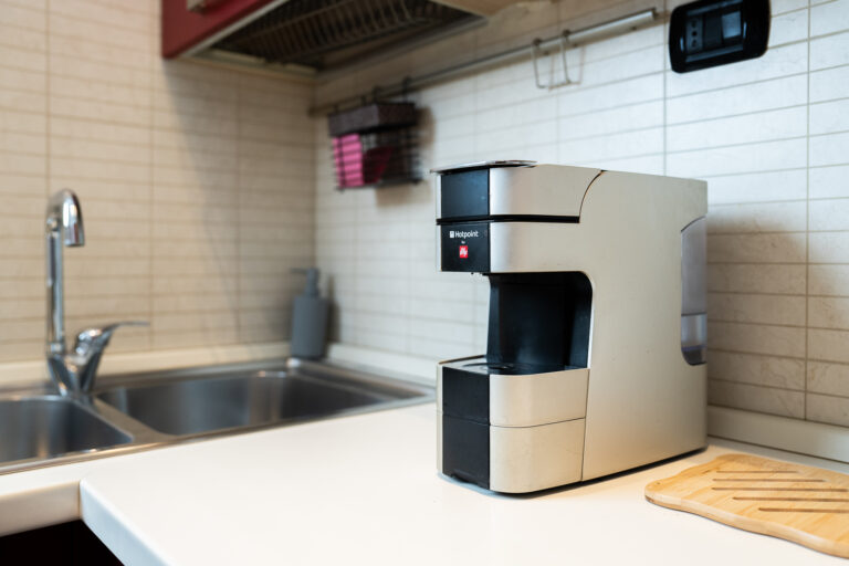 Cafetera plateada de diseño moderno en encimera blanca, junto a fregadero de acero inoxidable y tabla de cortar de madera.