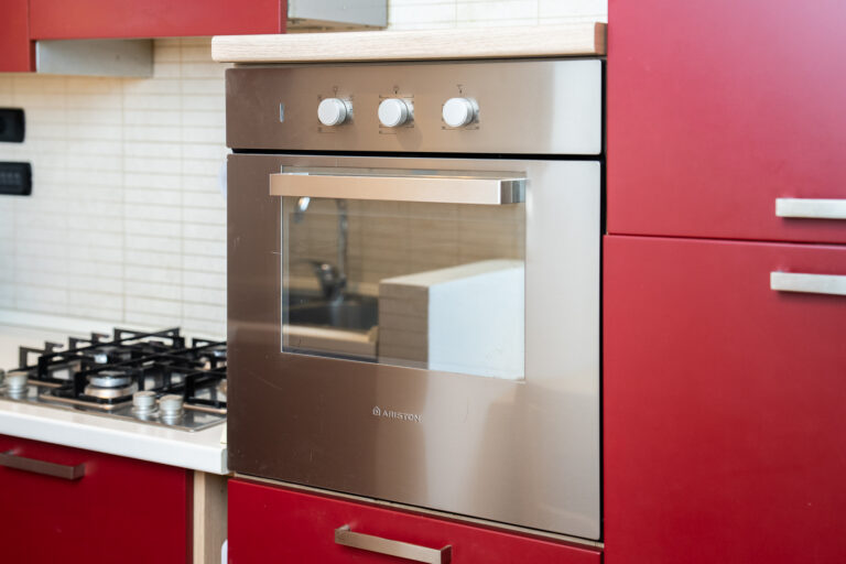 Horno de acero inoxidable con perillas, empotrado en cocina de gabinetes rojos, junto a una estufa de gas.
