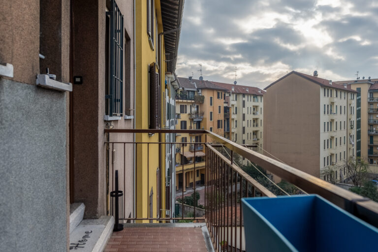 Vista desde un balcón con barandilla metálica, edificios coloridos, cielos nublados y antenas en los tejados.