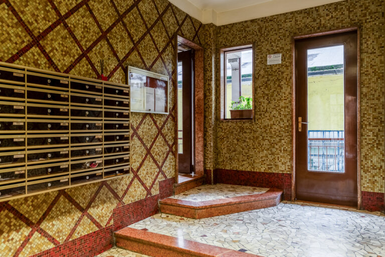 Vestíbulo con mosaicos marrones en las paredes, buzones metálicos, puerta de vidrio y ventana con planta en el alféizar.