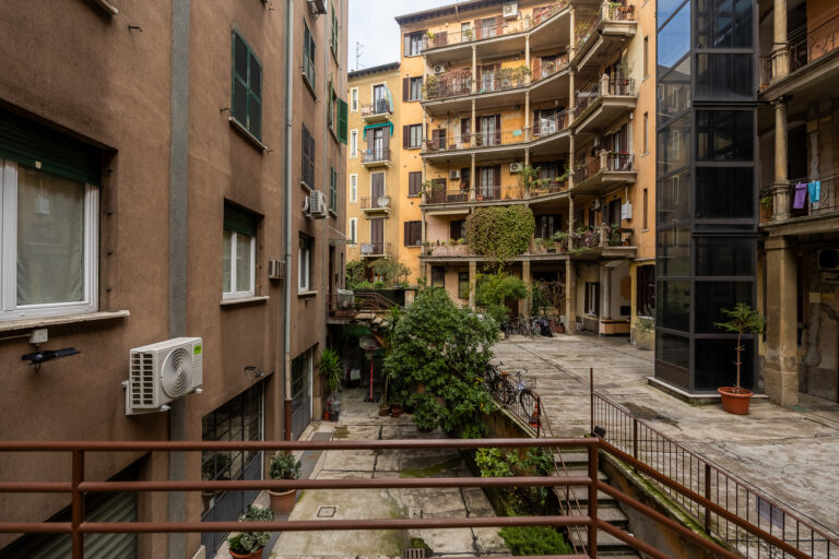 Edificio de apartamentos con balcones, plantas en el patio, bicicletas aparcadas y un ascensor exterior de cristal.