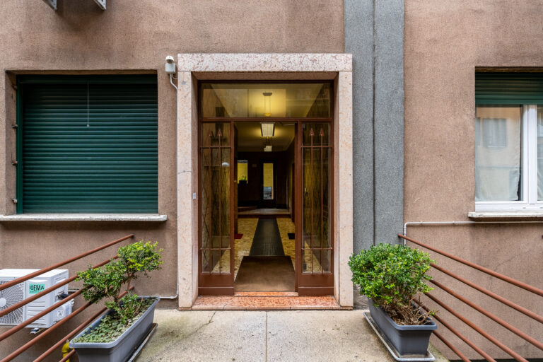 Entrada de edificio con puerta de cristal, marco de piedra y persianas verdes. Plantas en macetas junto a una barandilla metálica.
