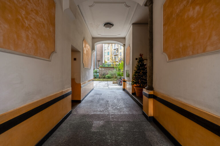 Pasillo con paredes pintadas de amarillo y beige, suelo de piedra gris, árbol de Navidad decorado, bicicletas y vegetación al fondo.