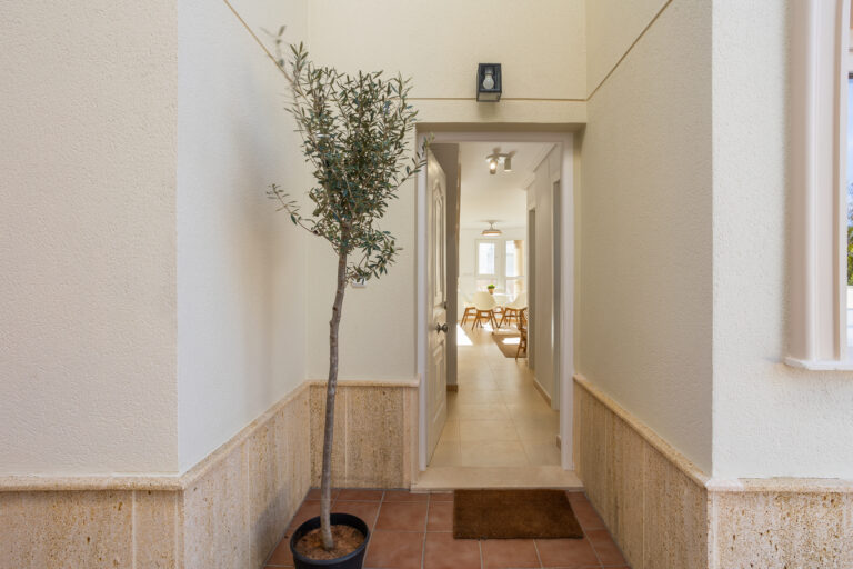 Entrada de puerta con maceta, pasillo hacia comedor con sillas blancas y mesa de madera. Paredes claras y luz natural.