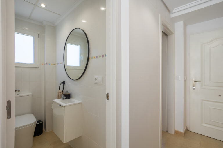 Baño pequeño con azulejos blancos, espejo circular, lavabo pequeño, inodoro blanco, ventana con luz natural.