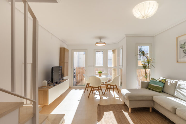 Sala de estar luminosa con sofá gris, mesa redonda blanca y sillas. Plantas y decoración moderna. Ventanales amplios y suelo claro.