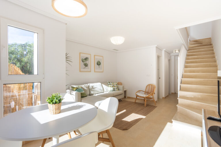 Sala luminosa con sofá claro, mesa redonda blanca, planta decorativa y escaleras beige. Luz natural entra por la ventana.