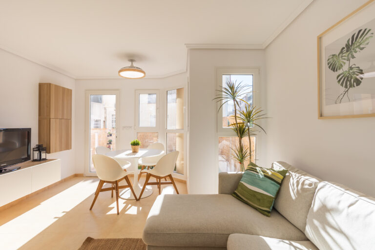 Sala de estar luminosa con sofá beige, cojín verde, mesa blanca y sillas, planta en esquina y ventanas con luz natural. Decoración sencilla.