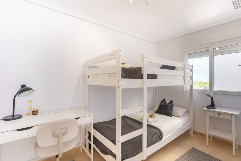 Habitación con literas blancas, escritorio con lámpara negra, silla beige, ventana grande y mesa auxiliar pequeña.