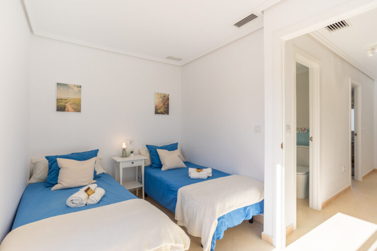 Habitación con dos camas individuales, ropa de cama azul, cojines beige, mesa de noche blanca y dos cuadros en la pared.