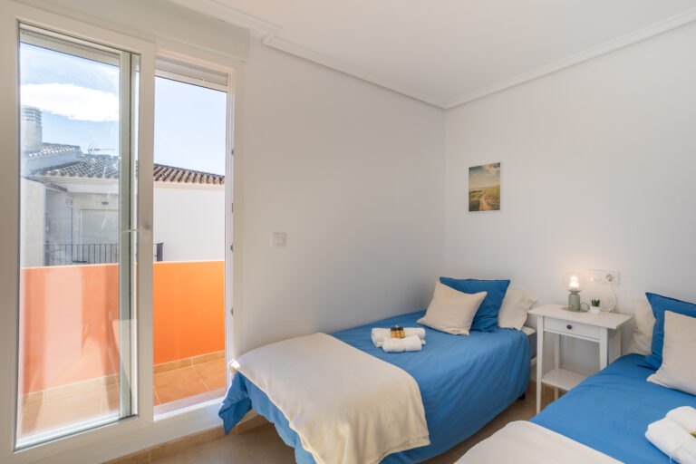 Habitación luminosa con dos camas individuales, ropa de cama azul, mesita blanca con lámpara, y un balcón exterior.