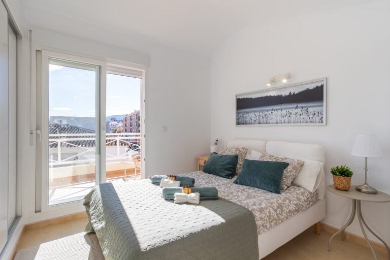 Dormitorio luminoso con cama doble, cojines verdes y beige, puerta de cristal hacia un balcón, y lámpara moderna sobre mesa auxiliar.