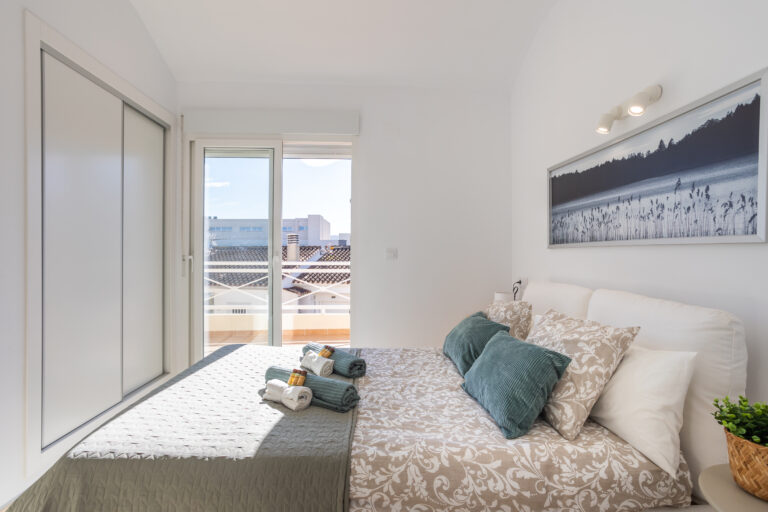 Dormitorio luminoso con cama doble, edredón estampado, cojines verdes, armario blanco, y puerta de cristal hacia un balcón.