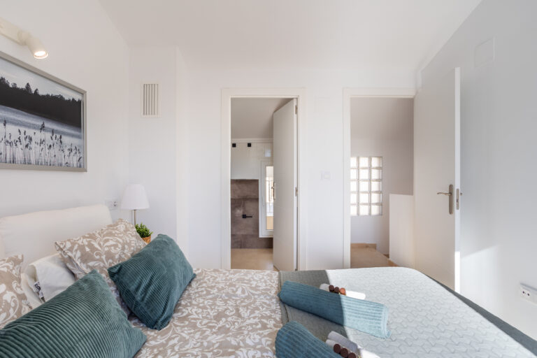 Dormitorio luminoso con cama doble, edredón floral y almohadas azules. Puertas abiertas hacia el baño y pasillo.