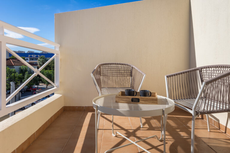 Terraza con dos sillas de mimbre, mesa redonda blanca con dos tazas negras. Barandilla blanca y vista urbana parcial.