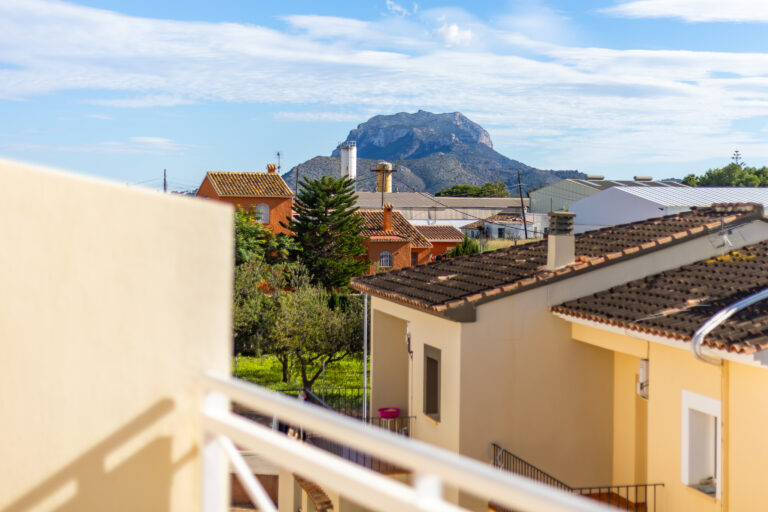 Casas con tejados marrones y naranja, árboles verdes en primer plano y una montaña rocosa al fondo bajo un cielo azul.