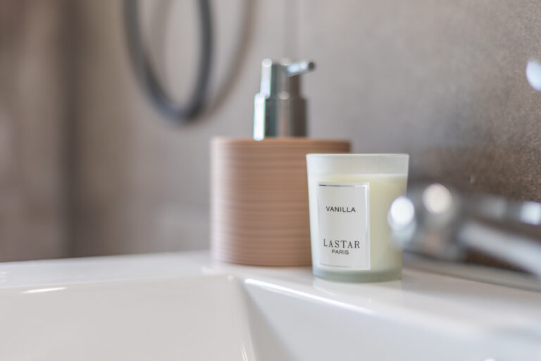 Vela de vainilla en frasco transparente junto a dispensador de jabón marrón sobre lavabo blanco. Fondo borroso.