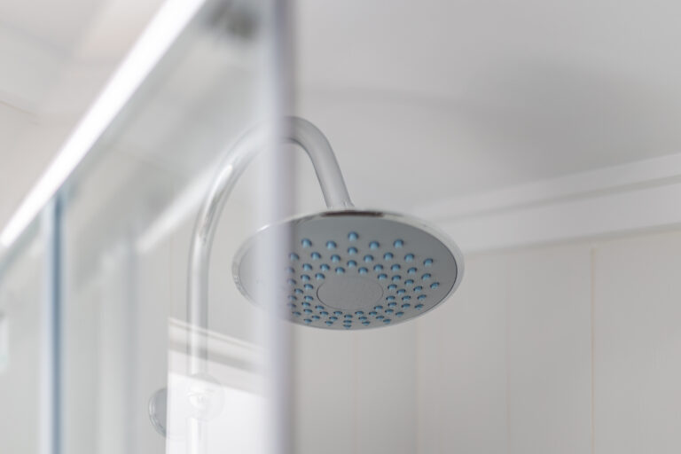 Ducha de cabeza redonda plateada con boquillas azules, parcialmente oculta por un panel de vidrio en un baño blanco.