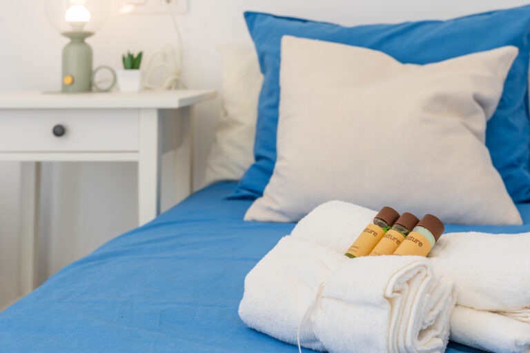 Cama con sábanas azules y almohadas beige, toallas blancas dobladas con botellas de baño. Mesa de noche con lámpara y planta.
