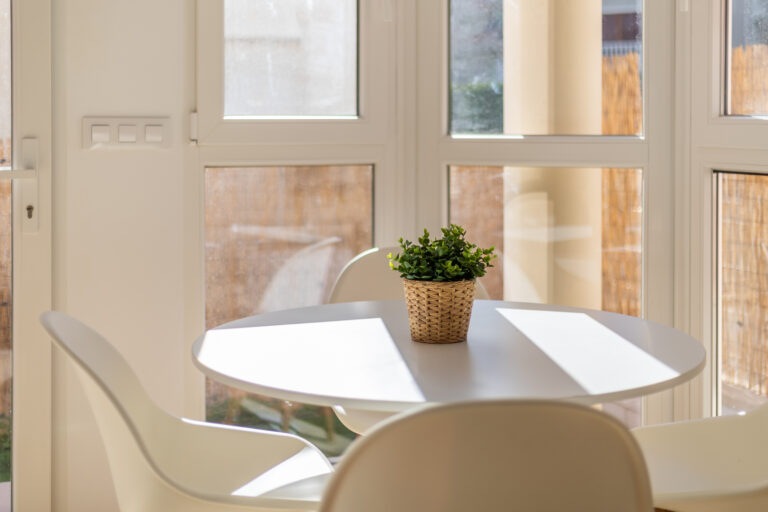 Mesa redonda blanca con sillas, planta en maceta tejida en el centro, luz natural entrando por ventanas grandes.