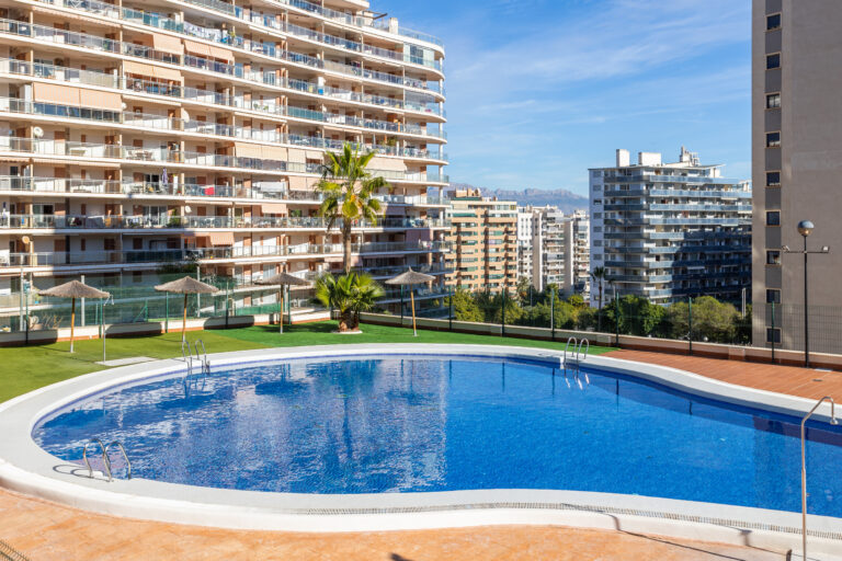 Piscina ovalada en un complejo de apartamentos altos con balcones, césped artificial y sombrillas de paja, bajo un cielo despejado.