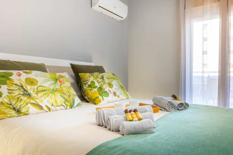 Habitación con cama doble, cojines verdes y estampados de hojas, toallas enrolladas, productos de tocador y ventana con cortinas.