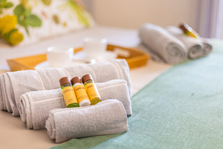Toallas blancas enrolladas con botellas de champú, crema y gel en cama. Fondo con tazas y bandeja de madera.