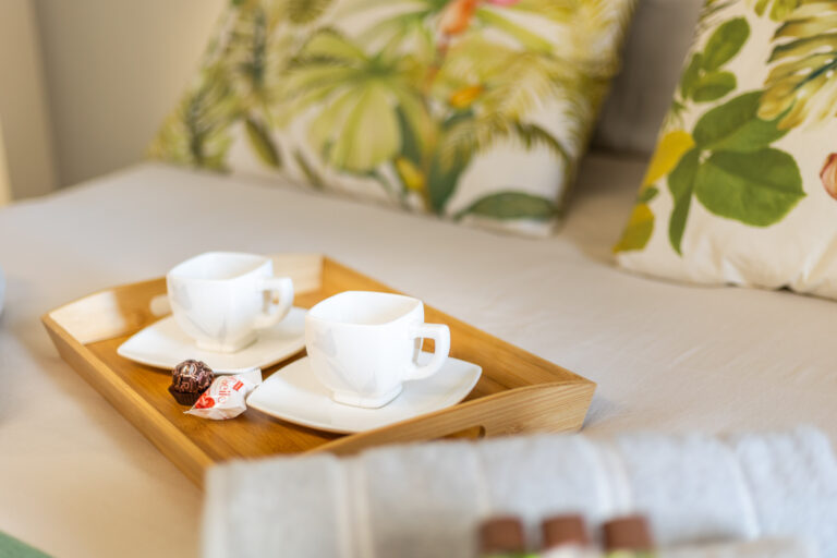 Taza de café blanca en bandeja de madera con caramelo, sobre cama con cojines de estampado tropical.