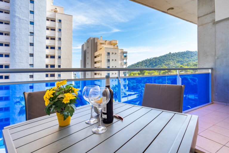 Balcón con mesa de madera, copas de vino, flores amarillas. Vista a edificios altos y colina verde bajo cielo azul.
