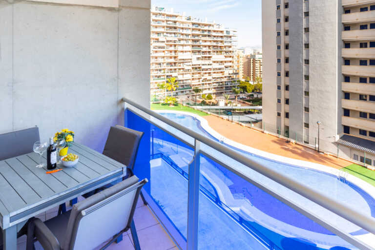 Terraza con mesa, sillas, botella de vino y flores. Vista de edificios y piscina azul al fondo.