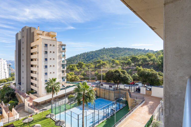 Edificio alto, cancha de pádel cercada, palmeras, colina verde al fondo, cielo azul claro con pocas nubes.