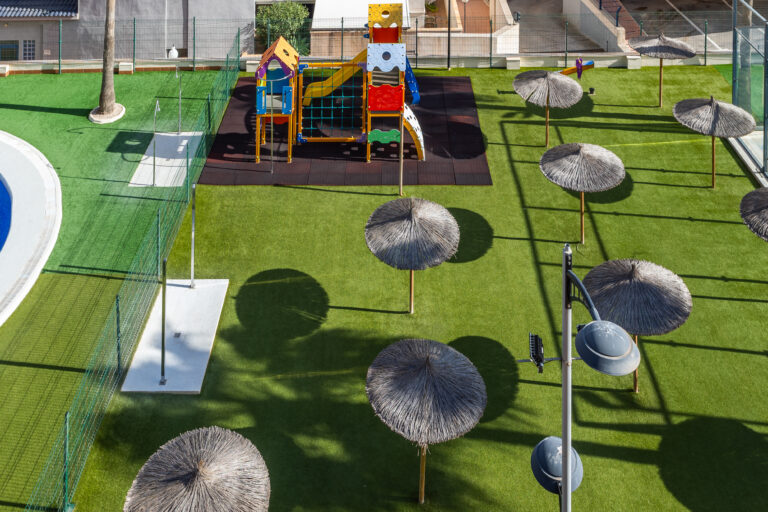Parque infantil con estructura de juego colorida, toboganes, red de escalada y sombrillas de estilo tropical sobre césped artificial.