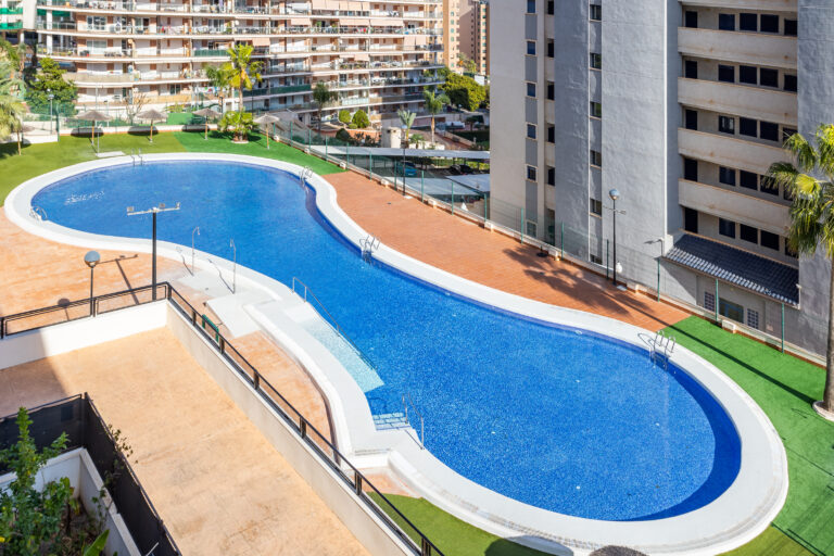 Piscina alargada en forma de riñón rodeada de edificios. Área pavimentada y césped artificial verde alrededor.