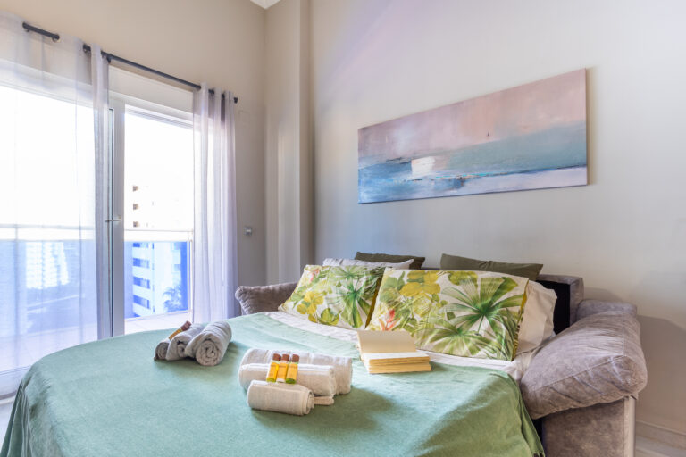 Cama con edredón verde, almohadas con estampado tropical, toallas y amenidades. Arte abstracto en la pared, iluminación natural.