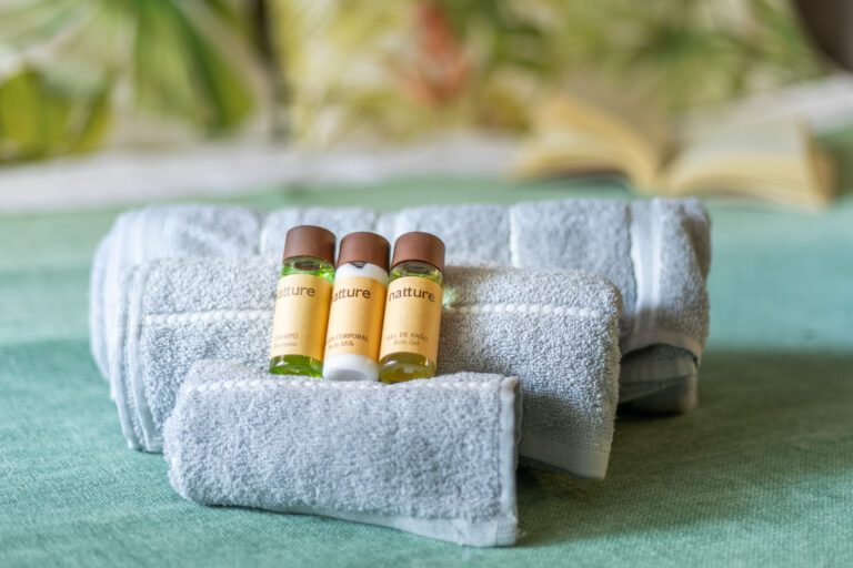 Tres botellas pequeñas de champú, leche corporal y gel de baño sobre toallas grises enrolladas. Fondo difuminado.