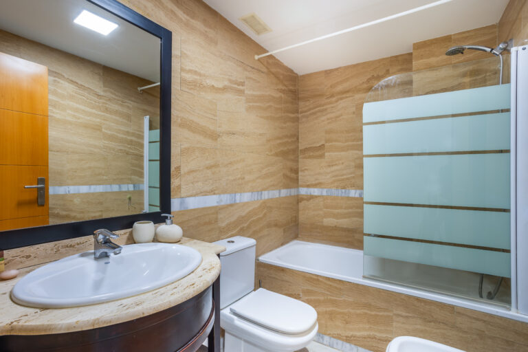 Baño moderno con lavamanos ovalado, espejo grande, ducha con mampara de vidrio esmerilado y paredes de mármol beige.