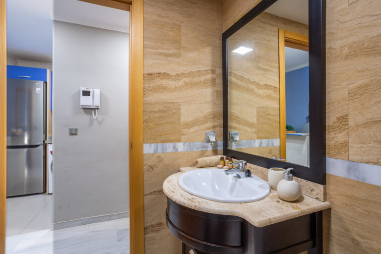 Baño con lavabo blanco, encimera de mármol beige, espejo grande y dispensador. Al fondo se ve una cocina moderna con nevera.