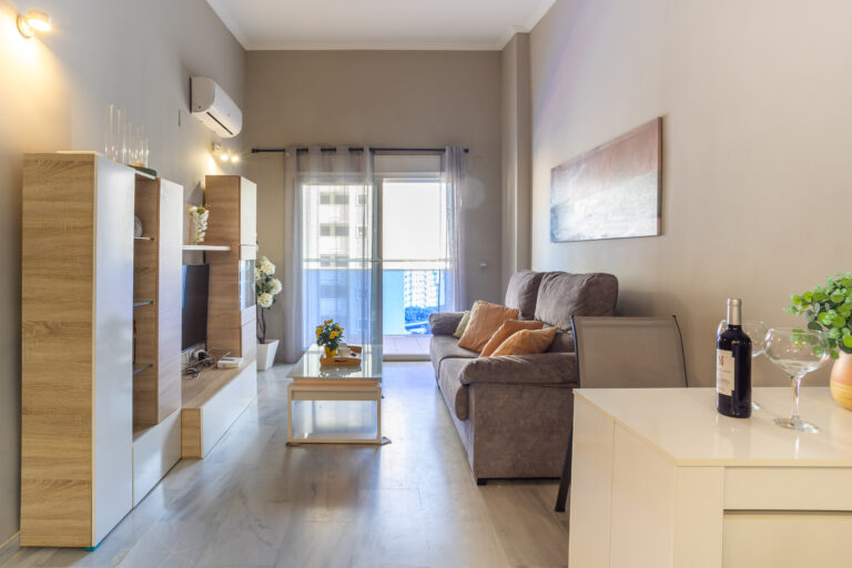 Sala moderna con sofá marrón, cojines anaranjados, mesa de centro con flores, estantería de madera y ventanal iluminado.