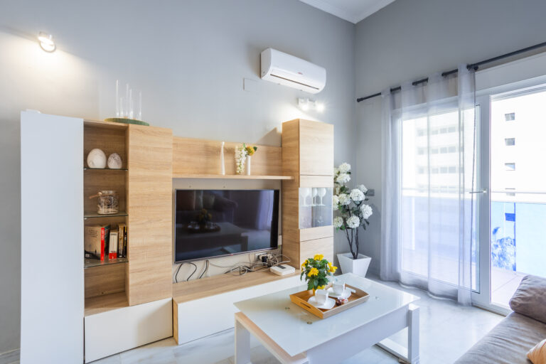 Sala moderna con paredes grises, mueble de madera clara, televisor, aire acondicionado, mesa de centro blanca y flores decorativas.