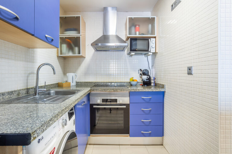 Cocina blanca con muebles azules, encimera de granito, horno, microondas, cafetera y frutas en un bol.