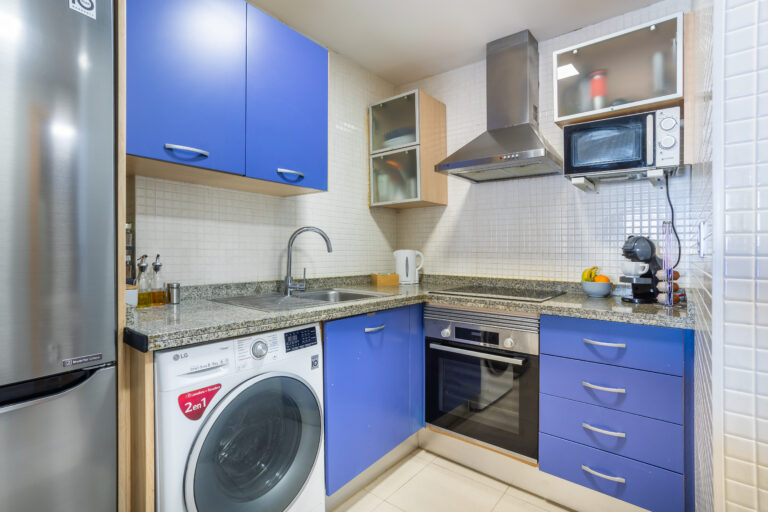 Cocina pequeña con gabinetes azules, electrodomésticos de acero, lavadora, horno, microondas y cafetera en encimeras de granito.